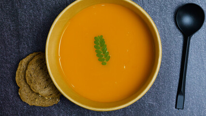 Creamy pumpkin soup served in a bowl, garnished with a green leaf, accompanied by two slices of bread and a black spoon, resting on a dark textured surface, presenting a warm and inviting meal