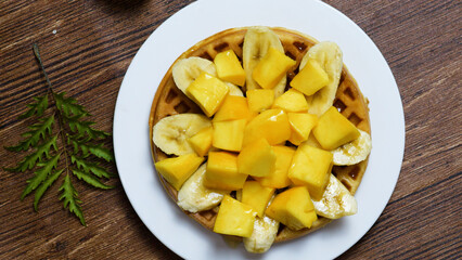 Round waffle topped with fresh banana and mango slices, creating a vibrant and appetizing breakfast or brunch presentation on a white plate