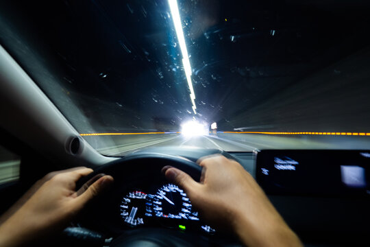 Hands gripping steering wheel while driving through illuminated tunnel at night, creating a sense of speed and motion