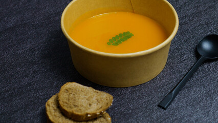 Warm, creamy carrot soup is served in a biodegradable bowl, accompanied by slices of whole wheat bread and a black spoon, creating a healthy and convenient meal option