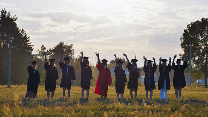 Happy graduates in graduation gowns waving at sunset in a field after ceremony