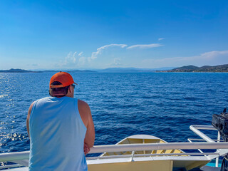 A man is looking out at the blue sea from the deck of a ferry on a sunny day with islands in the...