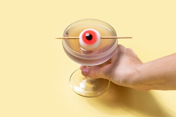 Hand holding purple Halloween cocktail garnished with candy eye on yellow background