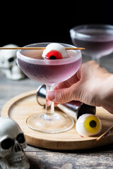 Hand holding purple Halloween cocktail garnished with candy eye on wooden table and black background. 