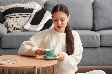 Beautiful young Asian woman with cup of hot tea on table in living room