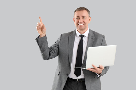 Portrait of mature businessman with modern laptop pointing at something on grey background - Powered by Adobe