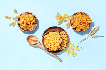 Wooden bowls with different types of raw pasta, spoon and wheat spikelets on blue background