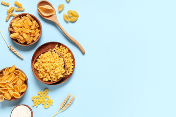 Wooden bowls with different types of raw tasty pasta, wooden spoon and spikelets on blue background