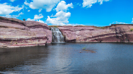 Image shows tranquil natural setting: calm pool with white waterfall from red cliffs, surrounded by reddish rock formations, blue sky, and scattered clouds.
