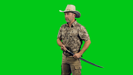 Fototapeta premium A middle aged stern man wearing camouflage with cowboy hat standing with sword against green screen