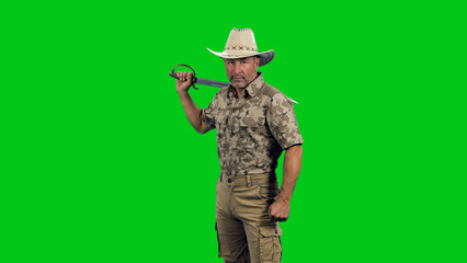 A middle aged rugged man wearing camouflage with cowboy hat standing with sword in hand and looking at camera against green screen