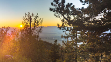 A serene mountain sunset with detailed tree silhouettes, golden-orange-blue skies, and a glowing sun casting warm light, evoking peace and natural beauty.