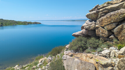 Tranquil lake under clear sky with rock formation and greenery, reflecting serene atmosphere of peaceful landscape view.