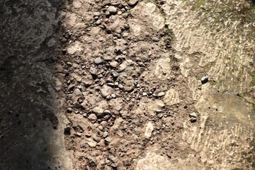 Close-up photo of a rough concrete surface with scattered gravel stones, dirt, cracks, and traces of green moss under sunlight. The image highlights raw urban texture, natural erosion.