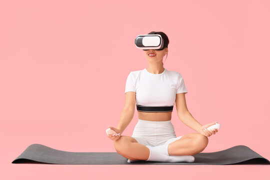 Sporty young woman in VR glasses with controllers meditating on yoga mat against pink background - Powered by Adobe