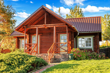 A rustic wooden log cabin with a tiled roof and stone foundation surrounded by greenery, flowers, and trees under a bright summer sky.