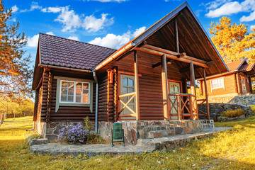 A rustic wooden log cabin with a tiled roof and stone foundation surrounded by greenery, flowers, and trees under a bright summer sky.
