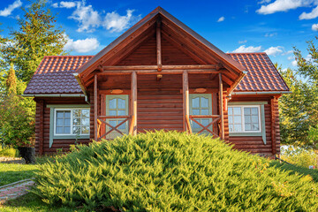 A rustic wooden log cabin with a tiled roof and stone foundation surrounded by greenery, flowers, and trees under a bright summer sky.