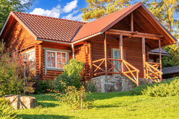A rustic wooden log cabin with a tiled roof and stone foundation surrounded by greenery, flowers, and trees under a bright summer sky.