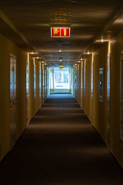 An empty hotel or apartment corridor with a long perspective, illuminated by ceiling lights, leading to a bright window at the end. The image has a mysterious, quiet atmosphere.