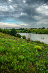 A stunning spring landscape with a calm river and a lush, green bank dotted with purple and yellow wildflowers under a dramatic and cloudy grey sky.