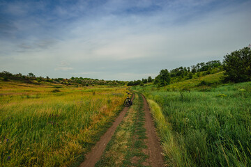 Obraz premium A mountain bike rests in the middle of a winding dirt road surrounded by lush green and golden fields, with rolling hills and a dense tree line in the distance under a cloudy sky.