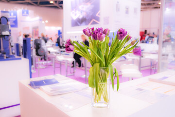 A vase of purple flowers sits on a table in a room with a lot of people
