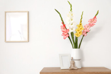 Vase with gladiolus flowers, statuette and blank frames on shelf near light wall