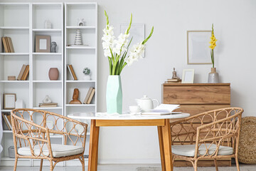 Vase with gladiolus flowers, coffee cup, teapot and book on dining table in room