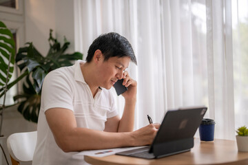 Focused asian man working from home, talking on phone and writing notes on tablet