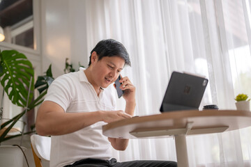 Asian freelancer talking on phone and working on tablet at home