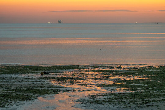 Luc-Sur-Mer, France - 08 08 2025: View of a ferry boat with the sunrise over the sea, reflections of colorful light and Le Havre far away at sunrise