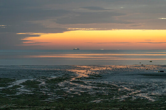 Luc-Sur-Mer, France - 08 08 2025: View of a ferry boat with the sunrise over the sea, reflections of colorful light and Le Havre far away at sunrise