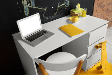 Children's workplace with blank laptop and books near chalk wall in room