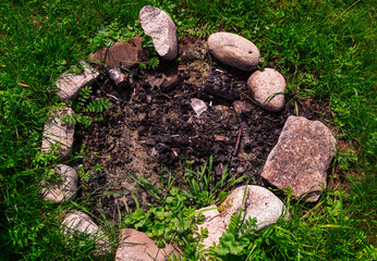 Charred remains of a campfire surrounded by rough stones on a patch of grass. The blackened wood and ashes evoke camping traditions, outdoor gatherings, and rustic countryside life.