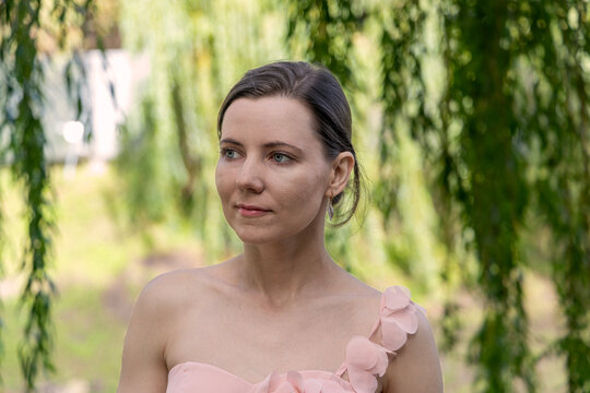 Elegant young woman in soft pink dress with floral details standing outdoors near willow tree branches in summer garden