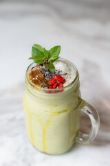Healthy avocado smoothie served in a mason jar, topped with shredded coconut, chia seeds, goji berries, honeycomb, and fresh mint leaves on marble background.
