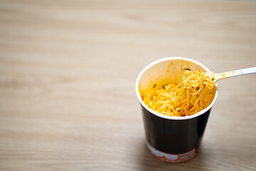 white disposable plastic fork with cooked instant noodles over blurred disposable paper cup with aluminum foil lid on wooden background. Eating behavior, effects of instant noodles on human health.