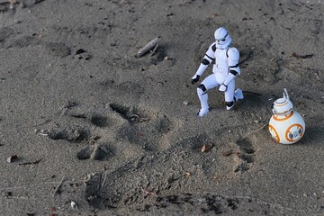 Fototapeta premium Figures of Star Wars clone trooper and ball shaped astromech droid BB8 examining tracks of a dog and little child in sand on shoreline of Danube river, southern Slovakia. 