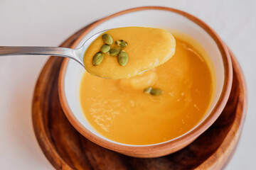 Spoon lifting creamy pumpkin soup from a bowl adorned with pepitas, served on a rustic wooden plate, inviting warmth for autumn meals