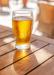 Glass of chilled beer on wooden table and blurred summer terrace at the background.