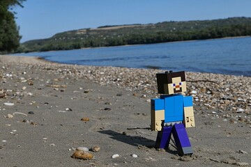 Obraz premium LEGO Minecraft action figure of smiling Steve walking on sandy bank of Danube river on borderline between Slovakia and Hungary. Forested opposite bank visible, blue skies, summer afternoon sunshine. 