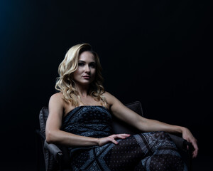 Young blond woman wearing elegant dress sitting on wooden chair in studio with dark background and...