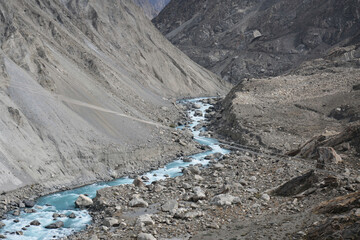 Turquoise mountain stream winding through a rocky, barren valley with steep slopes and scattered...