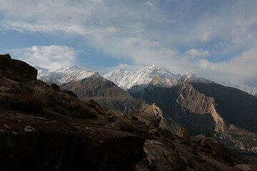 Snow-capped mountain range with rocky foreground beneath a cloudy blue sky.