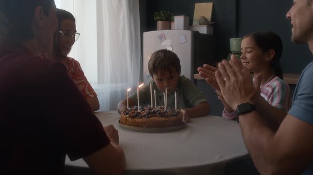 Little Caucasian boy blowing candles on homemade birthday cake, surrounded by loving parents, granny and little sister, all clapping hands and celebrating joyous moment together in cozy kitchen - Powered by Adobe