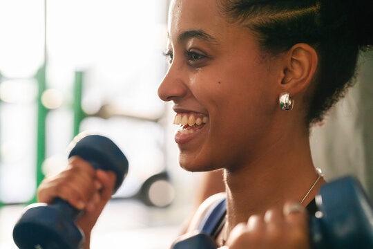 A happy young woman with dark skin lifts dumbbells in a gym. She is smiling and sweating, engaged in a focused and enjoyable strength training workout.