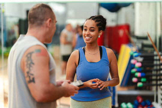 A man and a smiling young woman are conversing in a gym. He holds a phone while she listens attentively. They are engaged in a friendly discussion during their workout session.