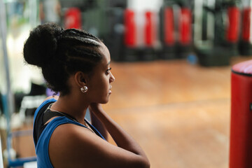 A young Black woman with braided hair styled in a high bun looks intently to the right. She wears a blue athletic top, observing the gym environment with red punching bags.