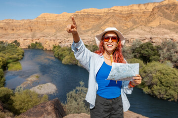 Female tourist holding a map and pointing while enjoying a sunny day by a river in a stunning...